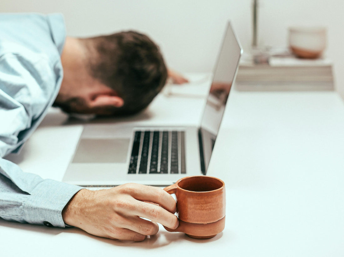 A man asleep face down on a desk beside a laptop and a half-finished coffee
